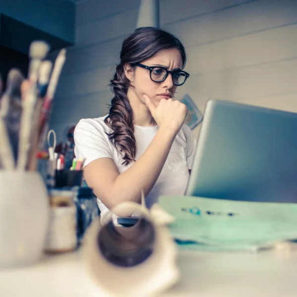 Focused young woman wearing glasses, working on a laptop in a creative studio, surrounded by art supplies — representing deep thought and strategy.